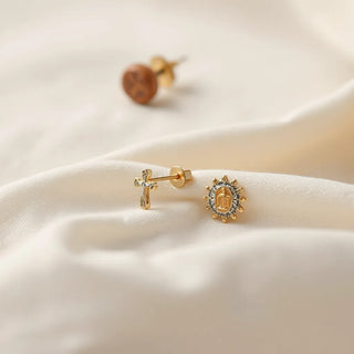 Elegant religious earrings with subtle cross and saint motifs on neutral fabric under soft window light